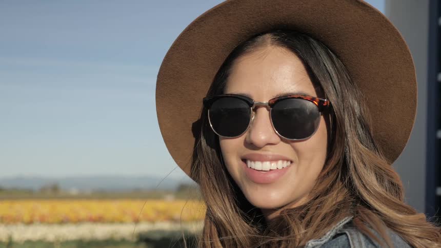 A woman in sunglasses and a wide brimmed hat smiles with rows of flowers in the background