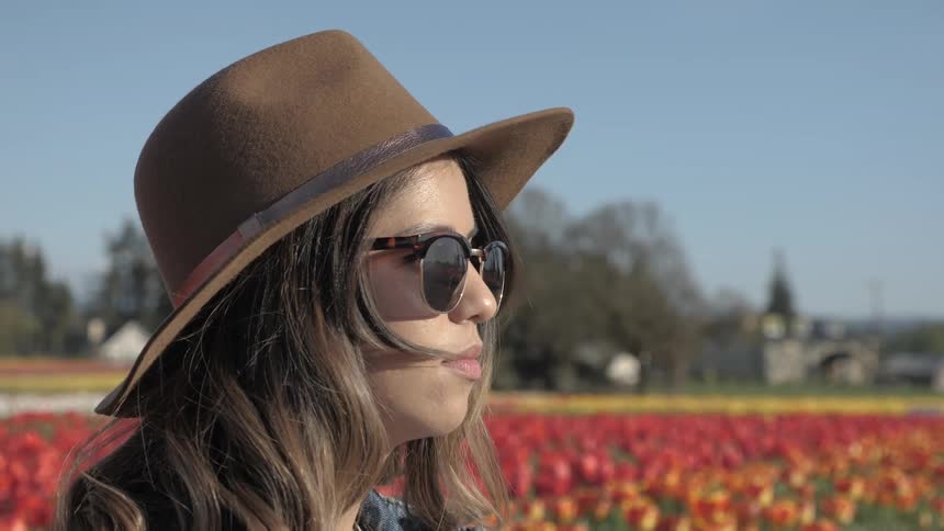 Dishwater blonde woman stands in the field of tulips wearing a brown leather hat and sunglasses looking straight ahead