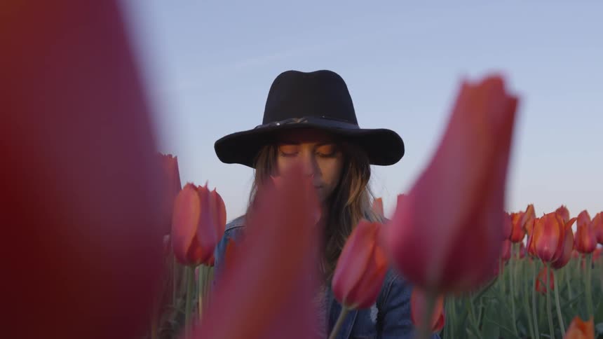 Woman wearing a black hat stands in a field of tulips as she smells one