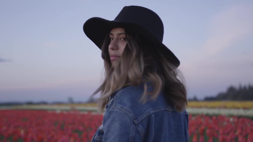 A female wearing a jean jacket and white brimmed hat walks through rows of red tulips and turns back to offer a partial smile
