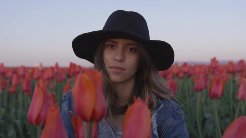 A woman in a black hat sits in a field of red tulips at sunset