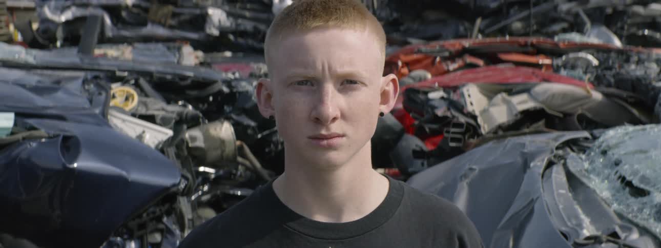 A teenager stares into the camera in a junkyard