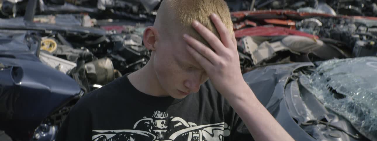 A young boy stands in a junkyard rubbing his head while looking down