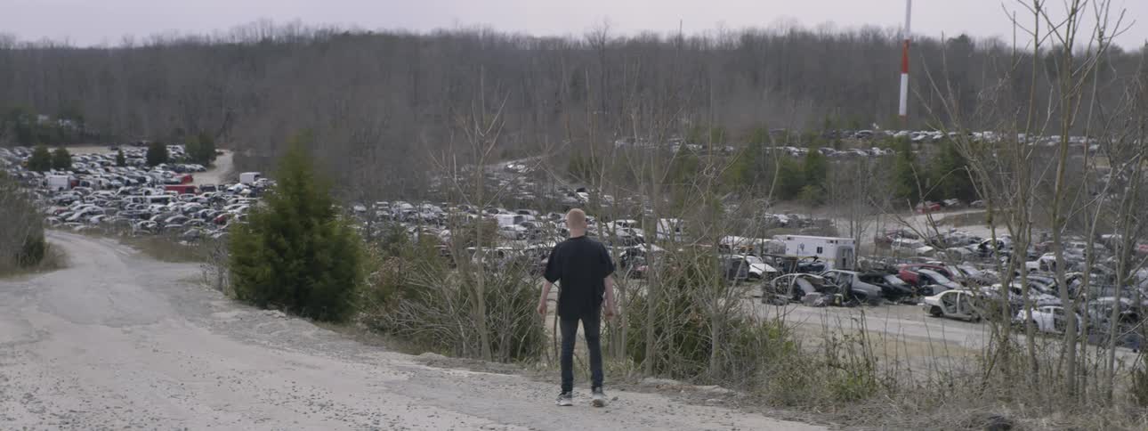 A teenager stands on the dirt road above a junkyard and throws a rock at the cars