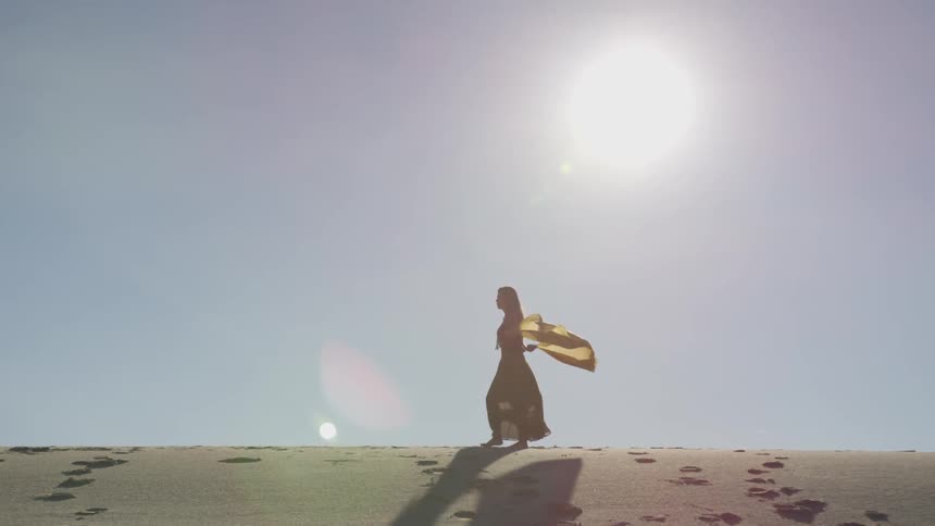 Woman with long yellow scarf walks to the left across the top of a sand dune in the desert