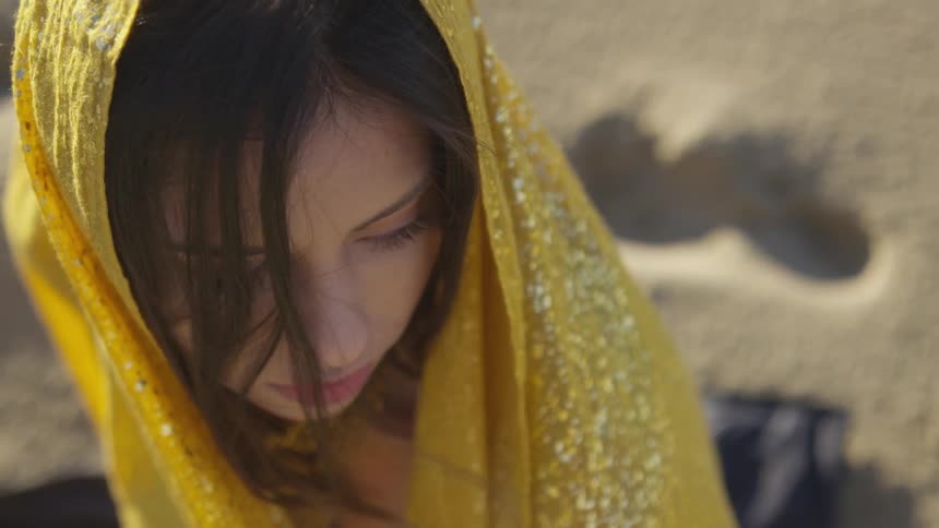 A woman wears a yellow and embellished scarf over her head as she sits outside on a sunny day