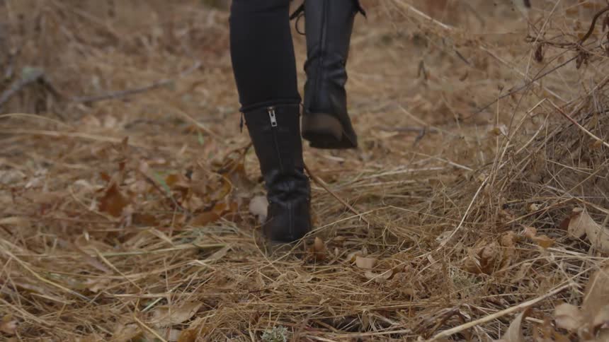 Woman wearing black pants and boots walks on faded grass in the daytime