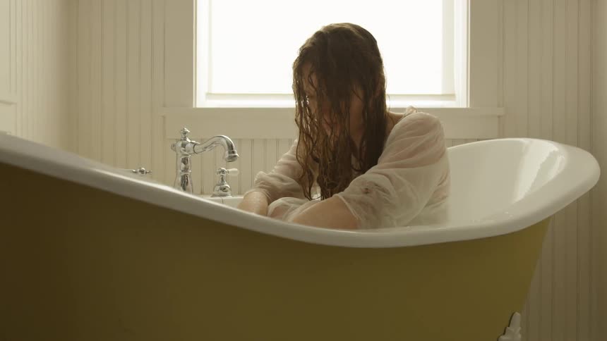 Woman sits in bathtub with wet clothing and hair looking down