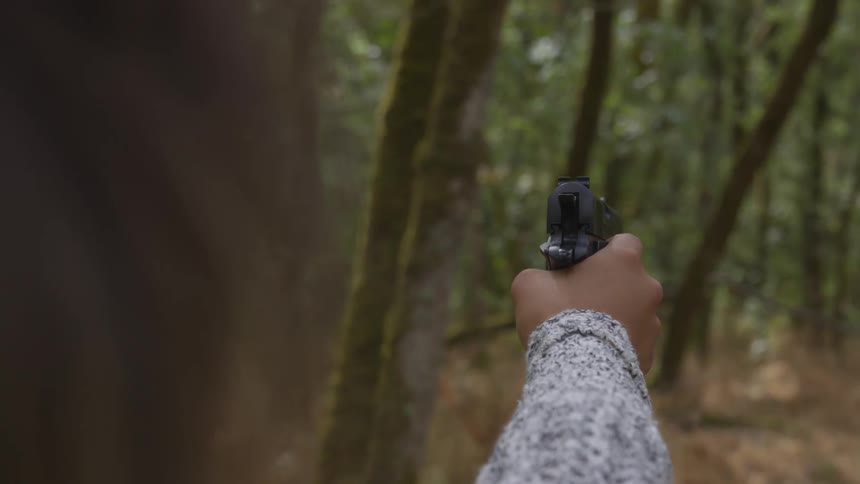 A woman points a handgun while standing in the forest