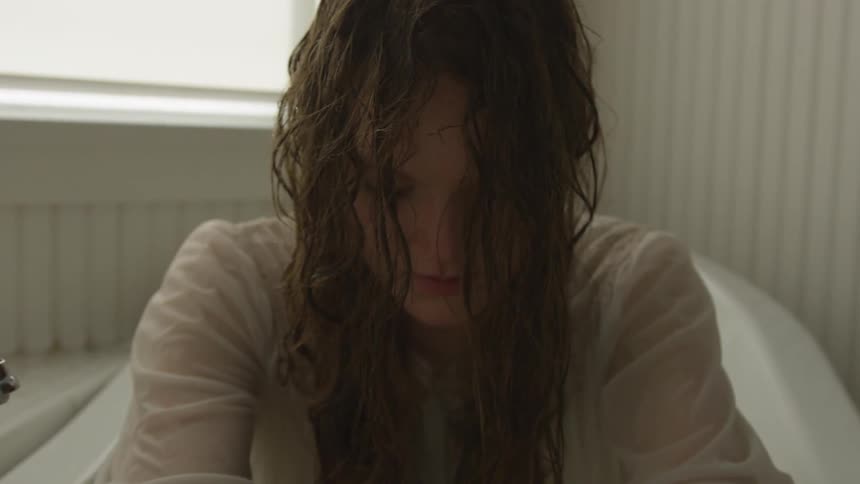 A brunette woman is looking down while sitting in a bathtub indoors