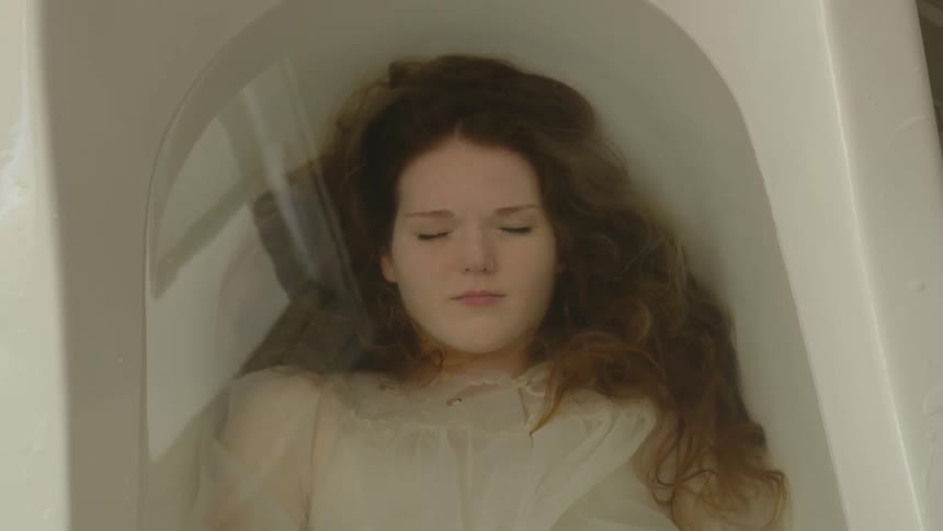 A young woman in white is laying submerged underwater in a bathtub with her eyes closed her blond hair swirling around her face