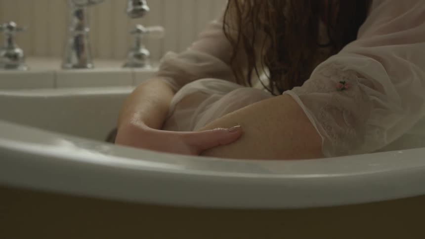 A clothed woman sits in a bathtub with her arms wrapped around her legs