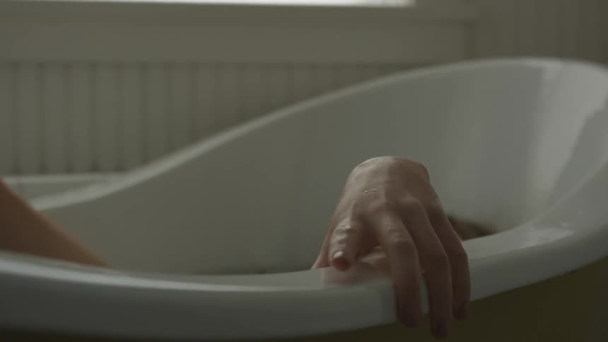 A white woman sinks down into a white bathtub while holding on to the edge of the tub