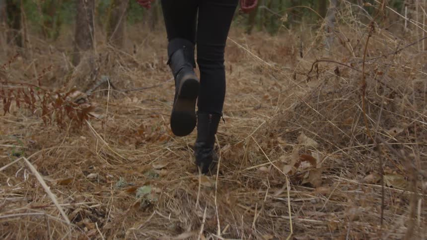 A woman wearing black boots and jeans walks down a path in the woods