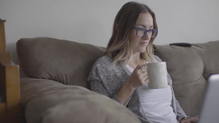 White woman sitting at the couch drinking coffee and using her laptop