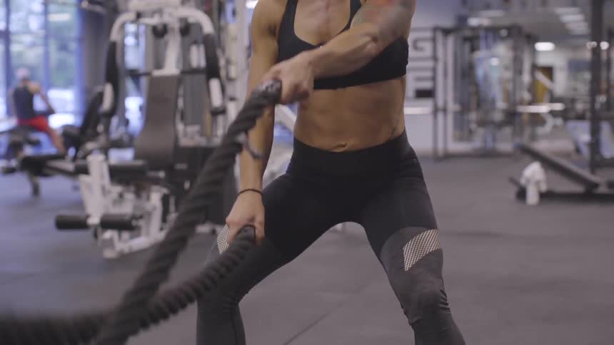 A blonde white female with a shoulder tattoo uses battling ropes in a gym