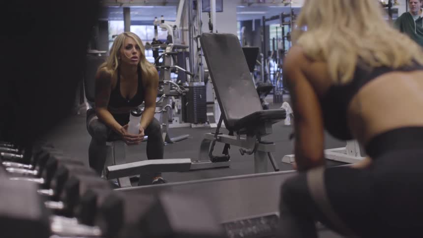 A white woman holds a plastic water bottle in her hand while sitting down at the gym