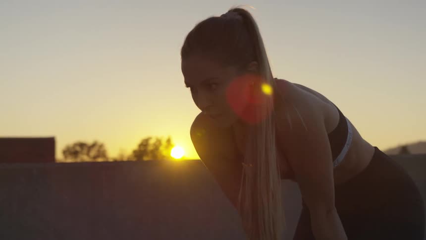 A white female in athletic apparel bends over with her hands on her thighs as he catches her breath with a sunset in the background