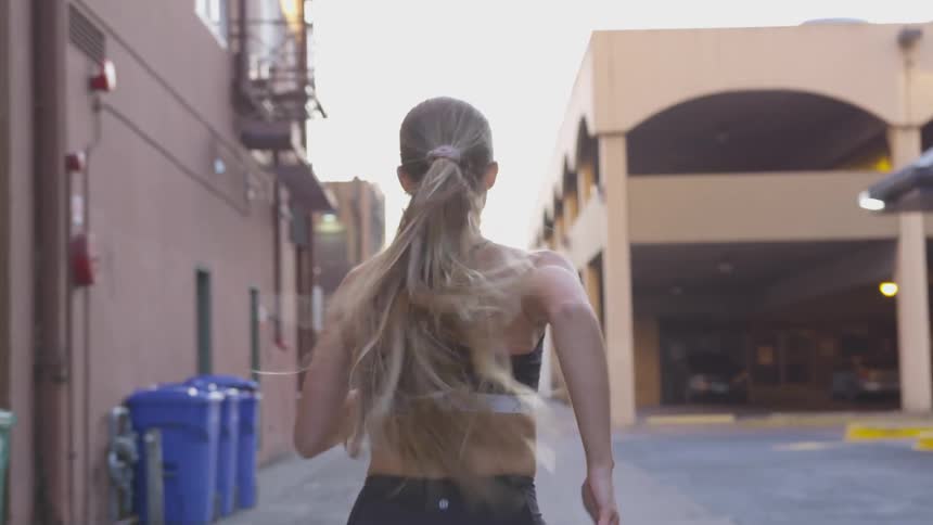A woman with her hair in a ponytail jogs past buildings in an alley