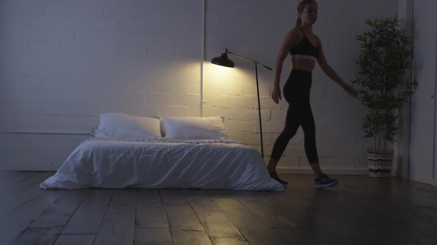 A white female in exercise apparel stands up from a mattress on a wood floor and crosses the room