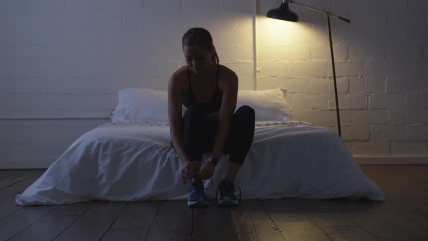 A woman ties her sneaker while sitting on a bed