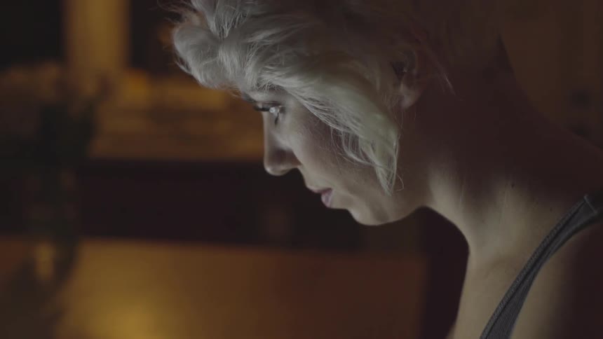 A woman looks down at her laptop screen while sitting in a dark room