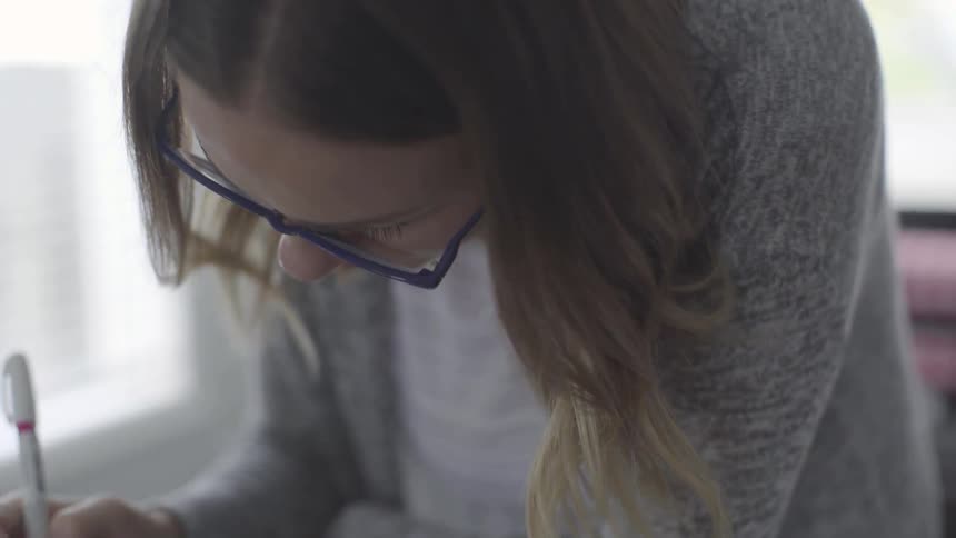 A young white woman with blue glasses writes something with a pen