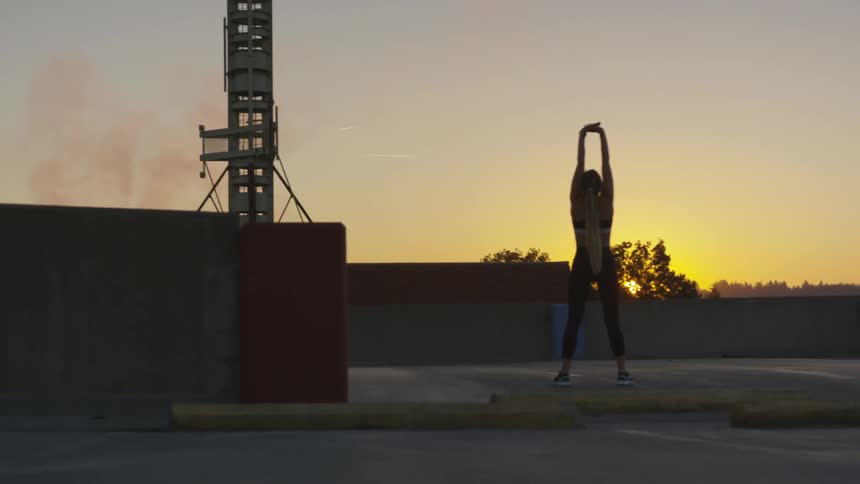 Woman performs warm up exercises on rooftop of building outside at dawn