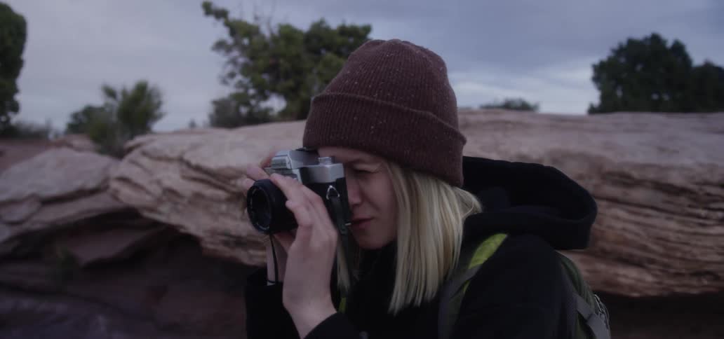 A white blonde woman wearing a coat and a knit hat takes a picture with a camera