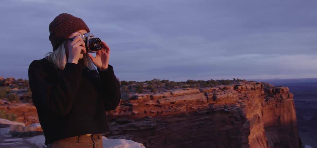 A woman takes pictures of a rocky canyon at sunrise while standing on a cliff