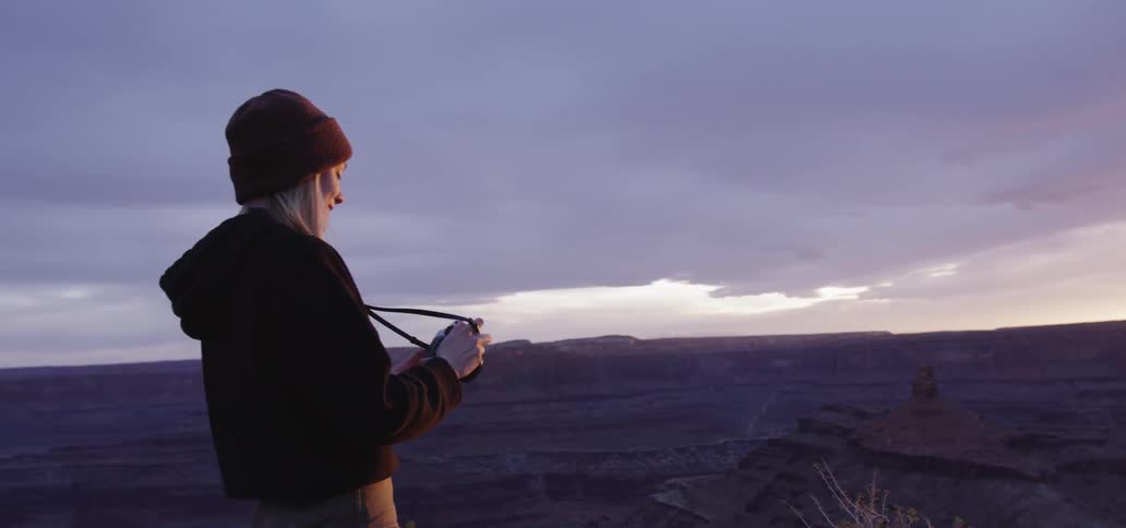 A young woman with blonde hair wears a jacket and a beanie as she stands atop the ridge of an expansive series of canyons and takes a picture with the sun very low in the sky