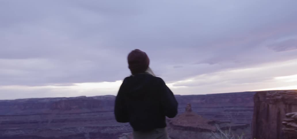 A young woman in a jacket and a knit beanie sprints to the edge of a cliff overlooking and enormous chain of canyons and pulls out her camera to get a photograph of the impressive landscape