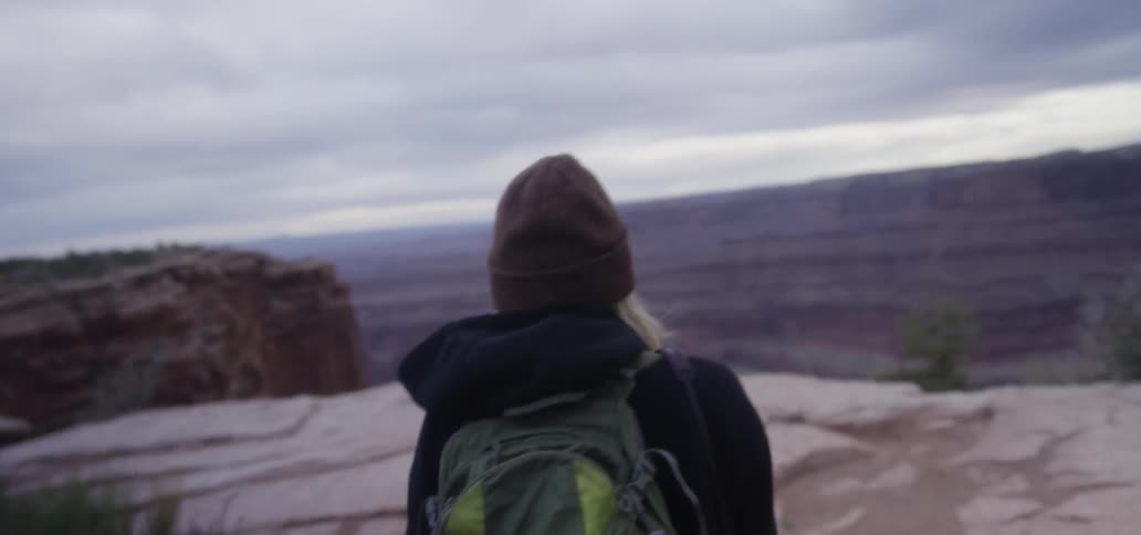 A woman wearing a backpack and a beanie walk toward the edge of a mountain top