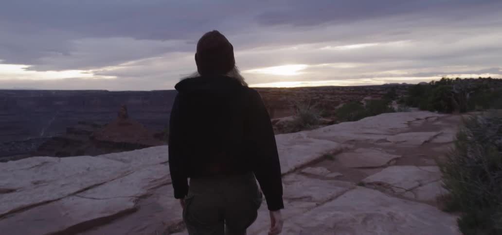 A white woman wearing a knit cap walks to the edge of a canyon rim