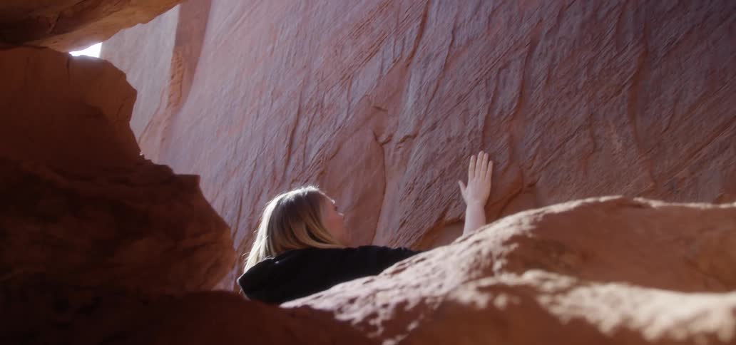 The woman is sitting on the red rock running her hands along it and feeling its texture
