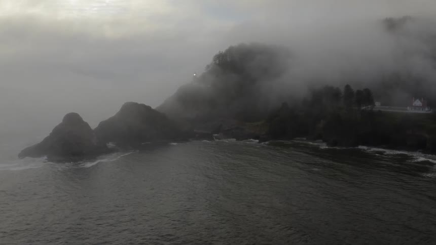 A lighthouse beacon flashes and rotates on a fog covered rocky shoreline