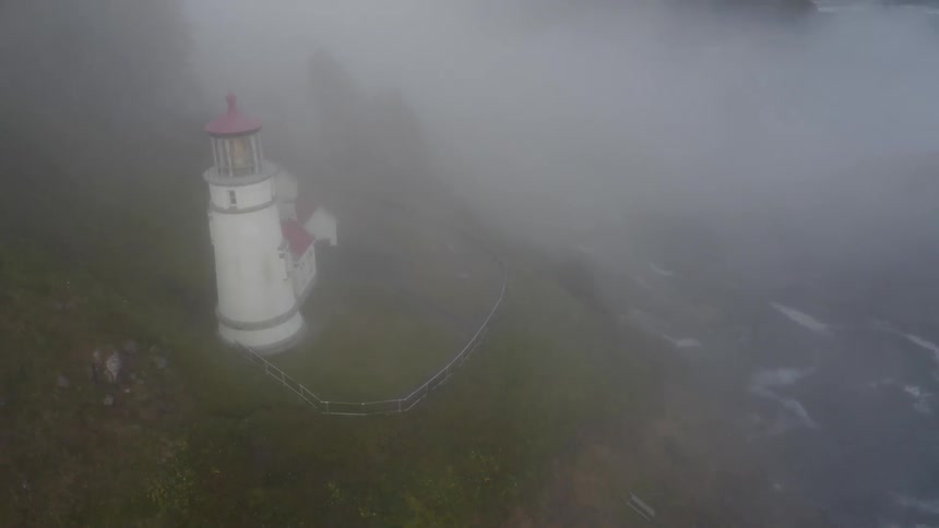 The camera looks down through clouds at a lighthouse next to the ocean