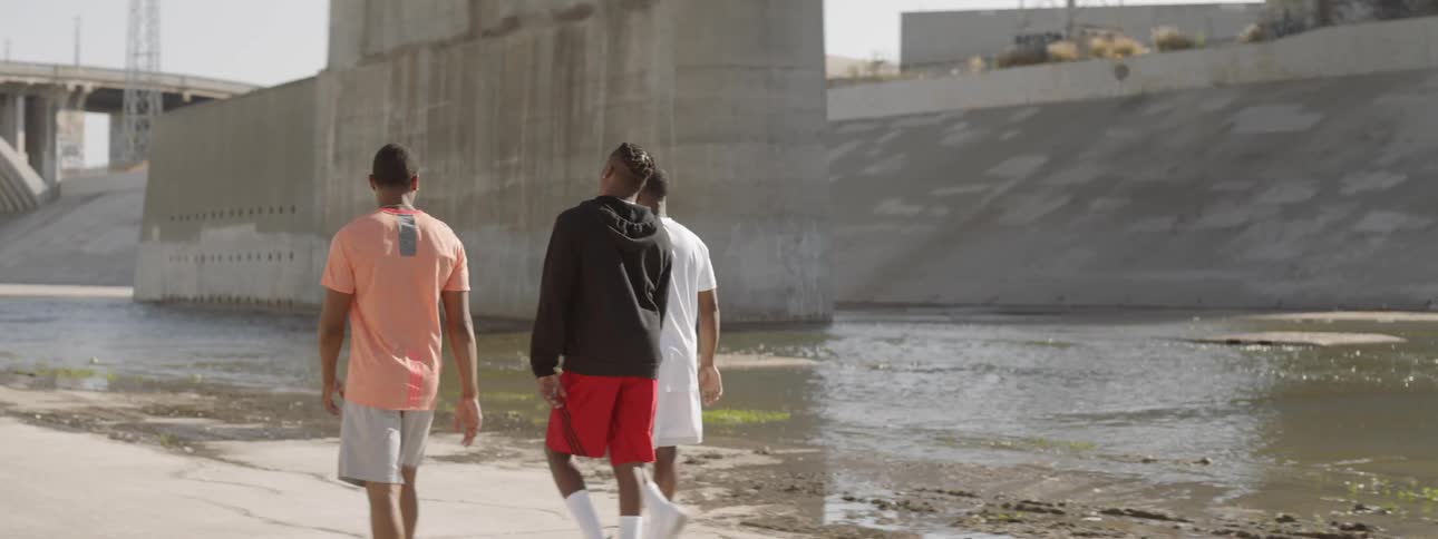 Three young black men walk outside on a sunny day in a moderately empty concrete rain water spillway with a small amount of water running through it