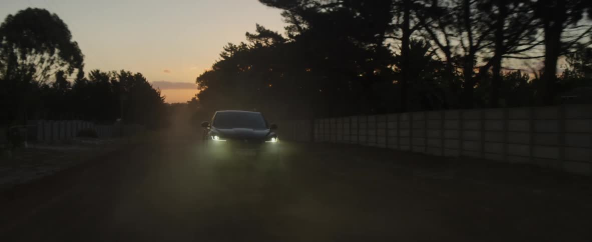 A car with its headlights turned on races along a dusty countryside road as the sun is low in the sky