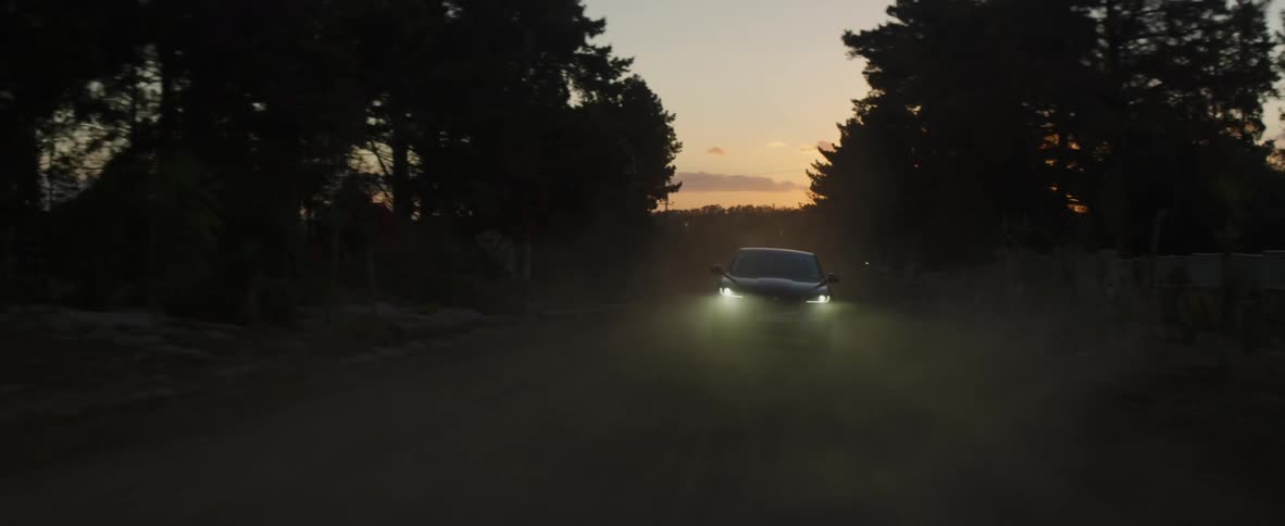 A car with the headlights on drives down a dirt road at dusk