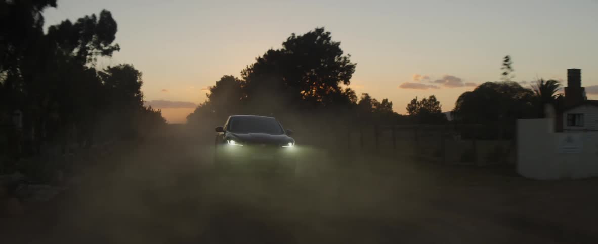 A car is speeding along a dusty road in a rural community as the sun is setting behind the trees
