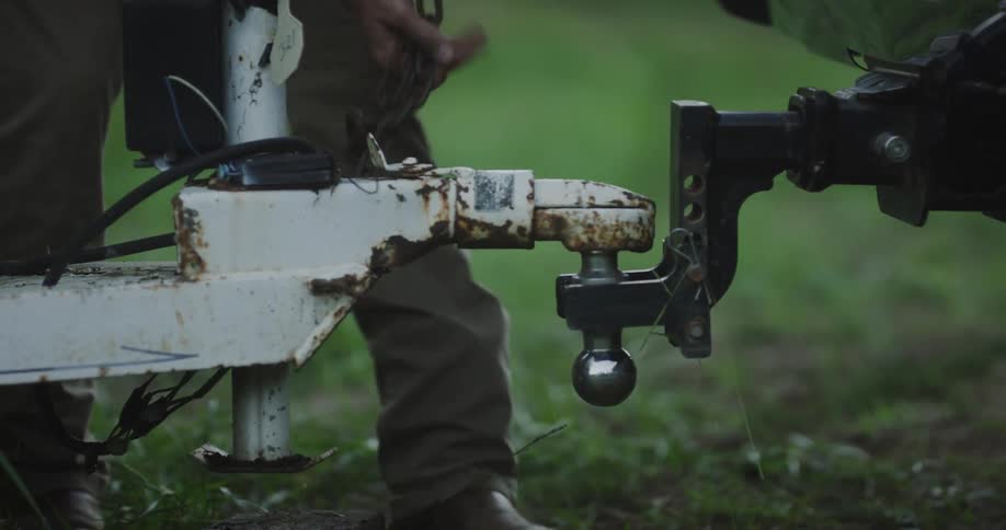 A man is preparing the connection between his truck and a trailer with a hitch covered in chipped white paint