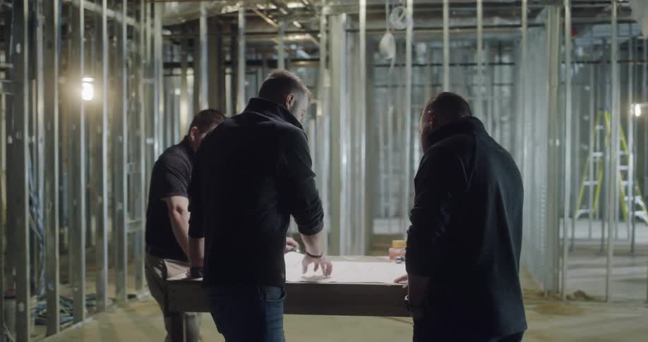 Three men discuss blueprints inside of a partially build structure