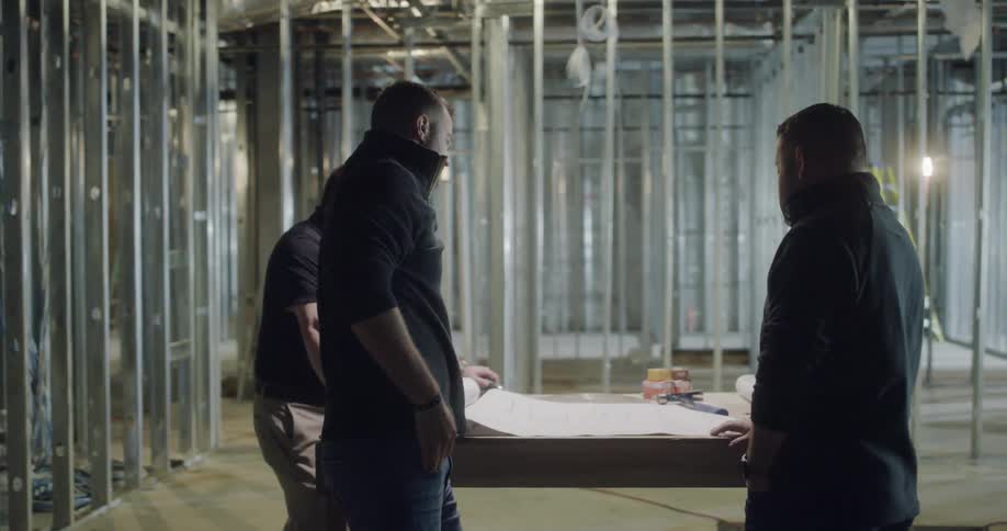 Three men stand by a table inside a steel framed building discussing blueprints and plans