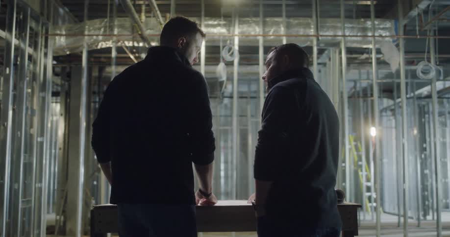 Two men in building under construction talk over plans at workbench