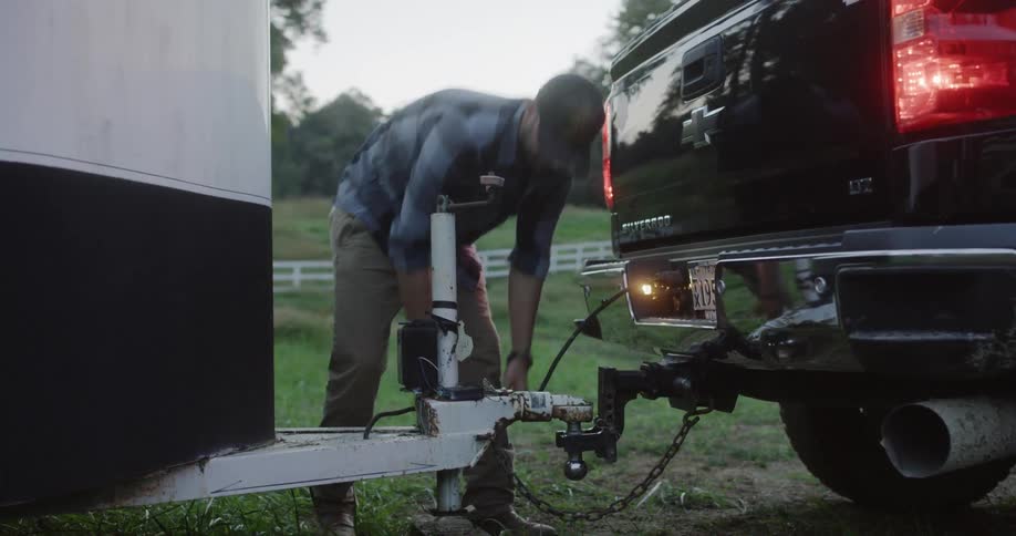 The man is in a grassy area hooking up his tow hitch and plugging in his lights on his truck