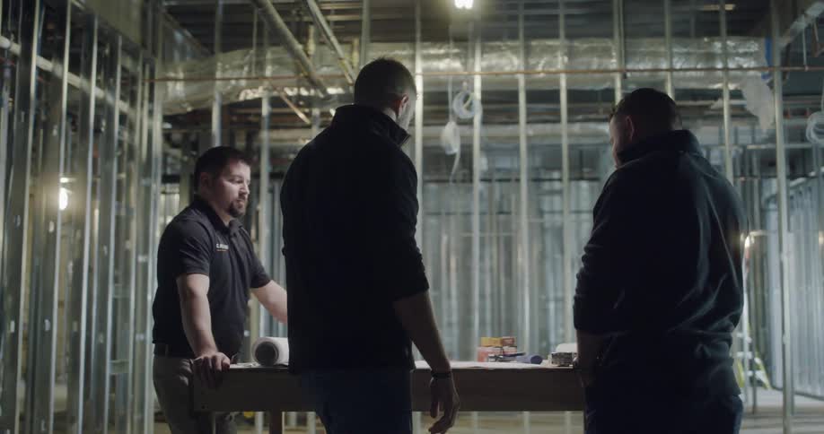 Three men stand around a table inside of the skeleton of a building that is under construction and they are discussing some of their plans