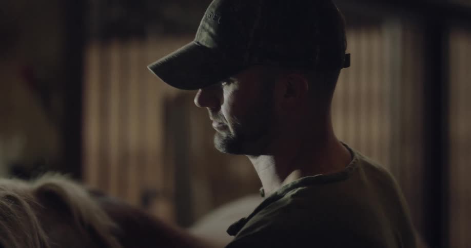 A man in a baseball hat is gentling brushing the hair of a horse inside of a barn as the horse is moving its head around