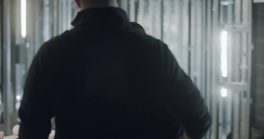 A man is standing amidst the scaffolding of a building that is under construction as he's looking over some building plans and a colleague walks up and gives him a pat on the shoulder