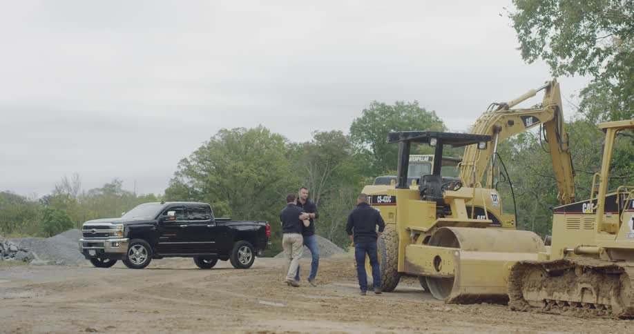 Three men greet each other at construction site with heavy machinery